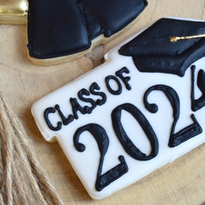 Pu&ograve; includere: Un biscotto bianco decorato con glassa nera, con il testo "Class of 2024" e un cappello di laurea nero con un fiocco dorato.