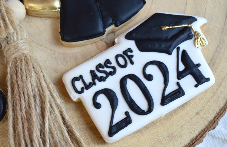 Pu&ograve; includere: Un biscotto bianco decorato con glassa nera, con il testo "Class of 2024" e un cappello di laurea nero con un fiocco dorato.