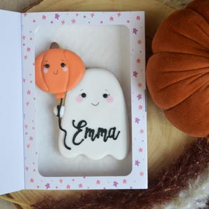 May include: A white ghost-shaped cookie with a smiling face and the name Emma written in black script, holding an orange pumpkin-shaped balloon, presented inside a white rectangular box with a pink star pattern border.