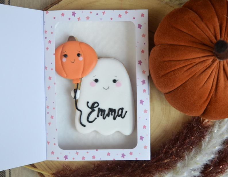 May include: A white ghost-shaped cookie with a smiling face and the name Emma written in black script, holding an orange pumpkin-shaped balloon, presented inside a white rectangular box with a pink star pattern border.