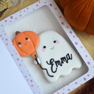 May include: A white ghost-shaped cookie with a smiling face and pink cheeks, holding an orange pumpkin-shaped balloon. The name Emma is written in black icing on the ghost. The cookie is inside a white box with a window and pink star patterns.
