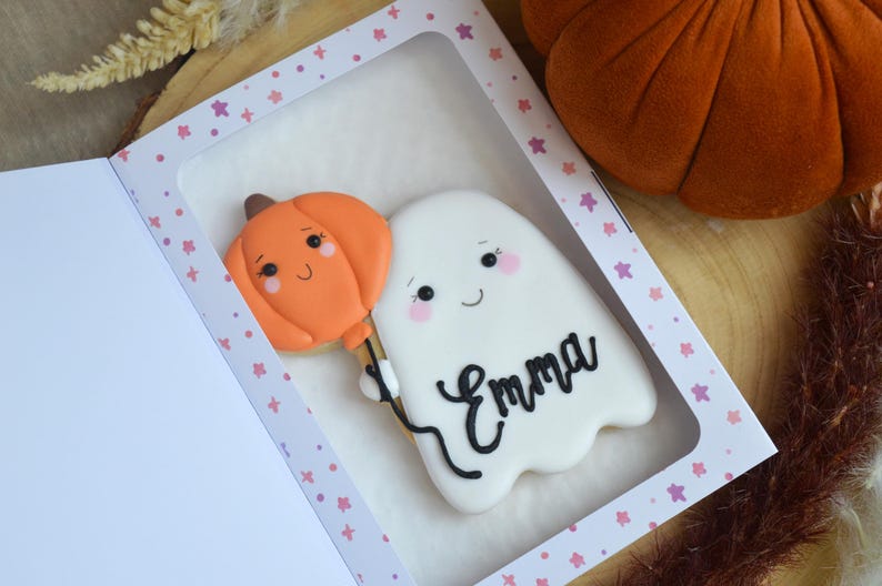 May include: A white ghost-shaped cookie with a smiling face and pink cheeks, holding an orange pumpkin-shaped balloon. The name Emma is written in black icing on the ghost. The cookie is inside a white box with a window and pink star patterns.
