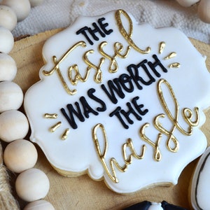 Pu&ograve; includere: Biscotto bianco decorato con glassa nera e lettere dorate. Il testo recita "The tassel was worth the hassle".