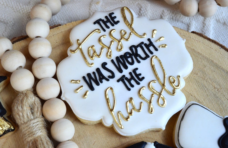 Pu&ograve; includere: Biscotto bianco decorato con glassa nera e lettere dorate. Il testo recita "The tassel was worth the hassle".