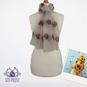 May include: A white scarf with a pattern of brown and white dog faces. The scarf is draped around a white mannequin. A separate image of a brown and white dog wearing a yellow and red hat and a yellow bow tie is in the bottom right corner.