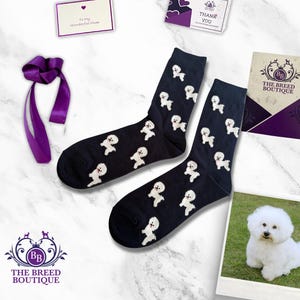 May include: A pair of black socks featuring a white Bichon Frise dog print. The socks are presented with a purple ribbon, a thank you card, and a card that reads "The Breed Boutique".