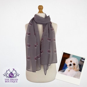 Puede incluir: Una bufanda gris con un estampado de perro blanco. La bufanda está en un maniquí blanco. El perro es de raza pequeña con pelaje blanco y ojos marrones. La bufanda está hecha de una tela suave y ligera.
