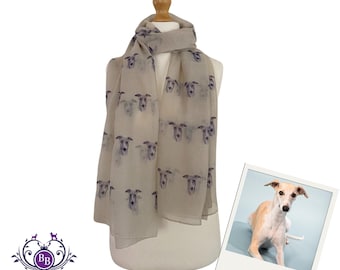 Puede incluir: Una bufanda beige claro con un patrón repetido de caras de perro whippet en azul oscuro. La bufanda está drapeada sobre un maniquí. Una foto enmarcada de un perro whippet está a la derecha. El logotipo de The Breed Boutique está a la izquierda.