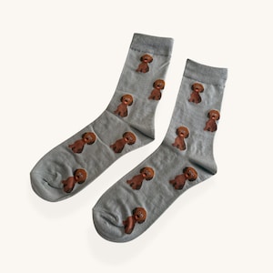 May include: A pair of light grey socks with a repeating pattern of brown poodle illustrations. The socks are made of a soft material, suitable for everyday wear. A fun and playful accessory.