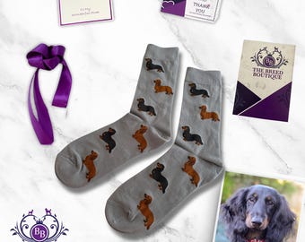 May include: A pair of gray socks featuring a repeating pattern of brown and black dachshund dogs. The socks are displayed with a purple ribbon, a card that says "To My Wonderful Mom", and a card that says "Thank You". A photo of a long-haired dachshund is also included.