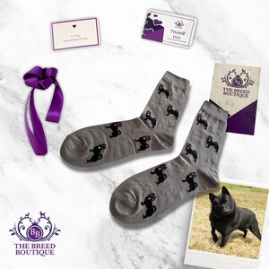 May include: A pair of gray socks with a repeating black dog pattern, alongside a photo of a black dog, a purple ribbon, and cards that say "Thank You" and "To My Wonderful Mom". The Breed Boutique logo is also visible.