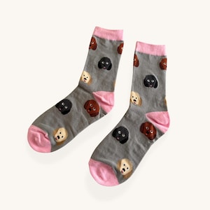 Puede incluir: Un par de calcetines grises con un patrón repetido de caras de perro en negro, marrón y crema. Los calcetines tienen talones y puños rosas. Los calcetines están hechos de un material suave.