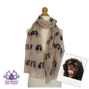 May include: A beige scarf with a repeating pattern of Cavalier King Charles Spaniel dog faces. The scarf is displayed on a mannequin. The Breed Boutique logo is visible in the lower left corner.