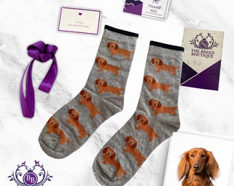 May include: A pair of gray socks with a repeating pattern of brown dachshund dogs. The socks have black cuffs. The image also includes a greeting card, a purple ribbon, and a small framed photo of a dachshund.