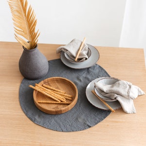 May include: A gray linen placemat with a wooden bowl filled with breadsticks. Two gray ceramic plates with linen napkins are on the placemat. A vase with a dried palm frond is in the background.