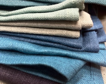 May include: A stack of folded linen napkins in various shades of blue, green, and gray. The napkins are arranged in a graduated color scheme, with the darkest blue at the bottom and the lightest green at the top.