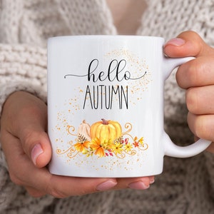 Puede incluir: Taza de cer&aacute;mica blanca con las palabras "hello AUTUMN" en letras negras cursivas y de imprenta. La taza presenta un dise&ntilde;o de acuarela de calabazas, follaje oto&ntilde;al y motas doradas. La taza es sostenida por una persona que lleva un su&eacute;ter color crema.