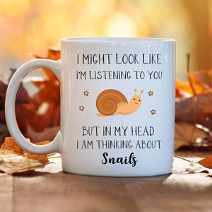 May include: White ceramic coffee mug with a black and white graphic of a snail and the text "I might look like I'm listening to you, but in my head I am thinking about snails".