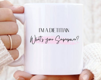 May include: White ceramic mug with black text that reads "I'm a Dietitian What's your Superpower?" with a pink watercolor accent.