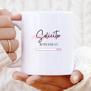 Può includere: Una tazza in ceramica bianca con un design rosa e nero che dice "Solicitor in Progress" con una barra di avanzamento sotto.