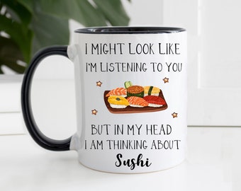 Könnte beinhalten: Weißer Keramikbecher mit schwarzem Henkel und Rand. Der Becher zeigt den Text "I MIGHT LOOK LIKE I'M LISTENING TO YOU BUT IN MY HEAD I AM THINKING ABOUT Sushi" zusammen mit einer Sushi-Illustration. Der Becher ist mit kleinen Sternen verziert.