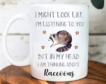 May include: White ceramic mug with a raccoon illustration and the text "I MIGHT LOOK LIKE I'M LISTENING TO YOU BUT IN MY HEAD I AM THINKING ABOUT Raccoons". The mug has a curved handle and is decorated with small gold stars.