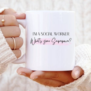 Könnte beinhalten: Weiße Keramiktasse mit schwarzem Text, der "Ich bin Sozialarbeiter. Was ist deine Superkraft?" lautet, mit einem rosa Aquarell-Akzent.