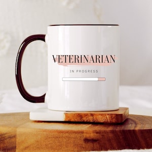 Puede incluir: Taza de cerámica blanca con asa y borde negros. La taza tiene un diseño rosa y negro que dice "Veterinario en progreso" con una barra de progreso.