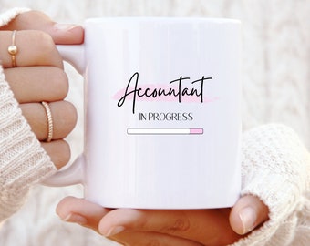 Op de afbeelding: Witte keramische mok met een roze en zwart ontwerp met de tekst "Accountant in progress" en een voortgangsbalk.