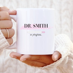 Puede incluir: Taza de cerámica blanca con un diseño rosa y negro. La taza dice "DR. SMITH in progress."