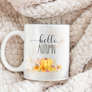 Puede incluir: Taza de cer&aacute;mica blanca con las palabras "hello AUTUMN" en escritura negra. La taza presenta un dise&ntilde;o de acuarela de calabazas y follaje oto&ntilde;al en tonos naranja, amarillo y marr&oacute;n. La taza tiene un asa grande y curvada.