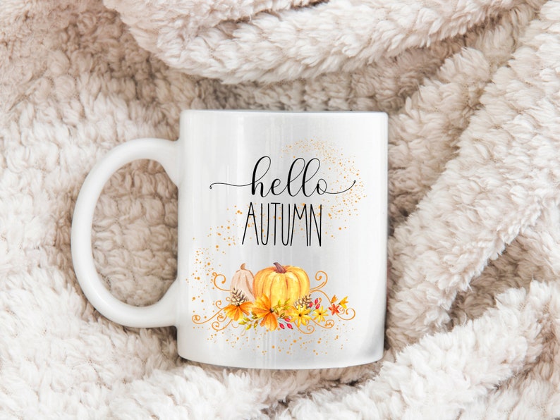 Puede incluir: Taza de cer&aacute;mica blanca con las palabras "hello AUTUMN" en escritura negra. La taza presenta un dise&ntilde;o de acuarela de calabazas y follaje oto&ntilde;al en tonos naranja, amarillo y marr&oacute;n. La taza tiene un asa grande y curvada.