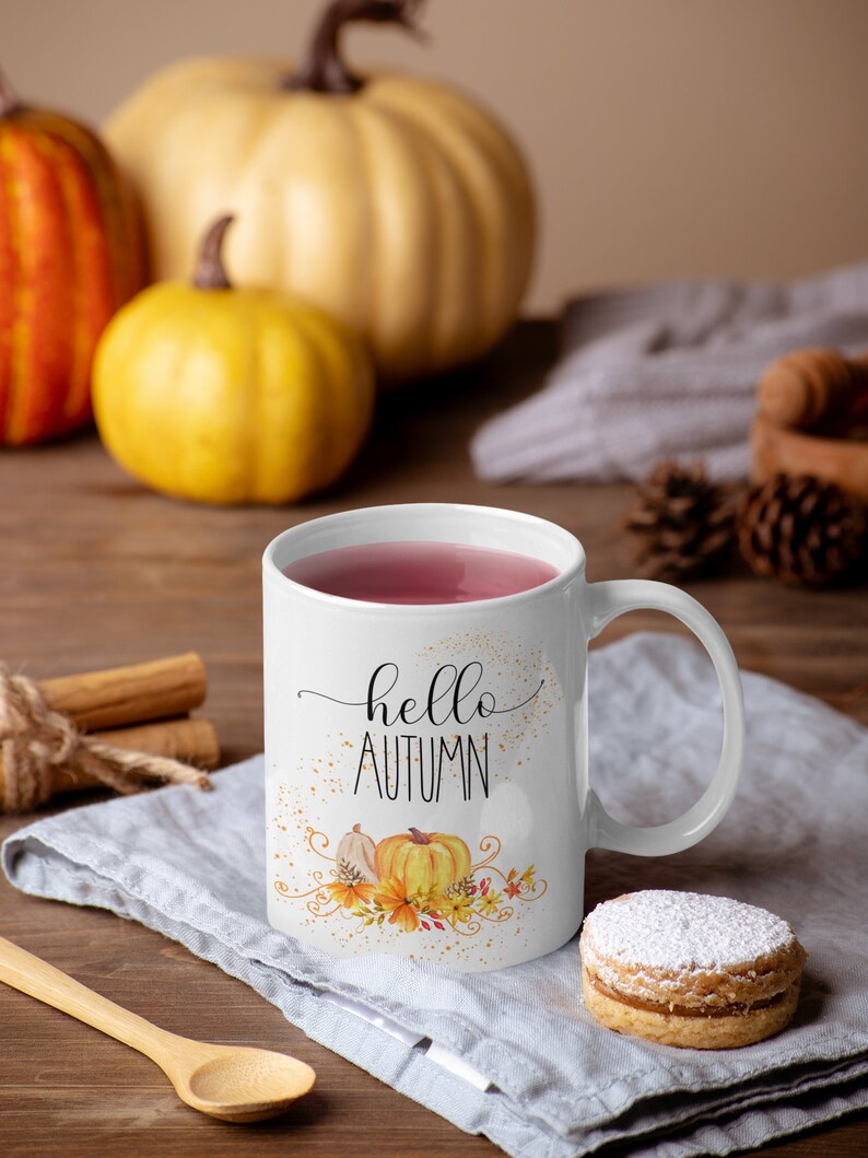Puede incluir: Taza de cer&aacute;mica blanca con las palabras "hello AUTUMN" en escritura negra, con un dise&ntilde;o de calabaza y flores. La taza est&aacute; llena de un l&iacute;quido rosa y se encuentra sobre un pa&ntilde;o gris. Calabazas y palitos de canela en el fondo.