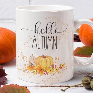 Puede incluir: Taza de cer&aacute;mica blanca con las palabras "hello AUTUMN" en letras negras cursivas y de bloque. La taza presenta un dise&ntilde;o de acuarela de calabazas y follaje oto&ntilde;al. Calabazas naranjas y hojas de oto&ntilde;o en el fondo.