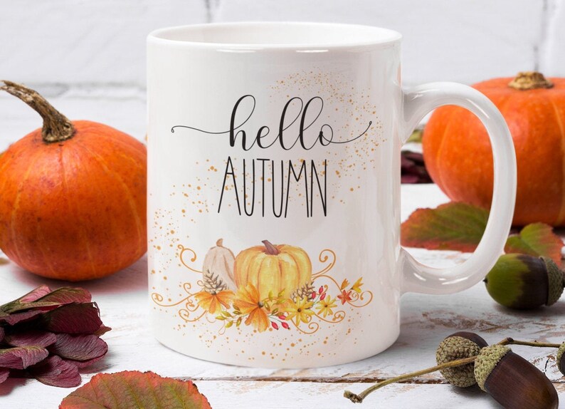 Puede incluir: Taza de cer&aacute;mica blanca con las palabras "hello AUTUMN" en letras negras cursivas y de bloque. La taza presenta un dise&ntilde;o de acuarela de calabazas y follaje oto&ntilde;al. Calabazas naranjas y hojas de oto&ntilde;o en el fondo.