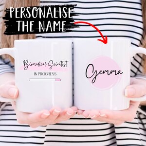May include: White ceramic mug with a pink circle on the right side that says "Gemma" in black cursive. The left side of the mug has the text "Biomedical Scientist" in black cursive with a pink progress bar underneath that says "In Progress".