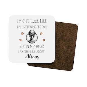 May include: A white coaster with a black and gray alien illustration and the text "I might look like I'm listening to you but in my head I am thinking about Aliens".