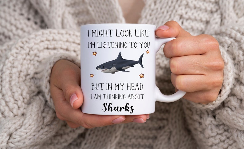 Pu&ograve; includere: Una tazza in ceramica bianca con un'illustrazione in bianco e nero di uno squalo e il testo "I might look like I'm listening to you but in my head I am thinking about Sharks".