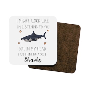 Pu&ograve; includere: Sottobicchiere bianco con un'illustrazione di squalo nero e grigio e il testo "I might look like I'm listening to you but in my head I am thinking about Sharks".