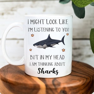 Pu&ograve; includere: Una tazza in ceramica bianca con un'illustrazione di squalo in bianco e nero e il testo "I might look like I'm listening to you but in my head I am thinking about Sharks".