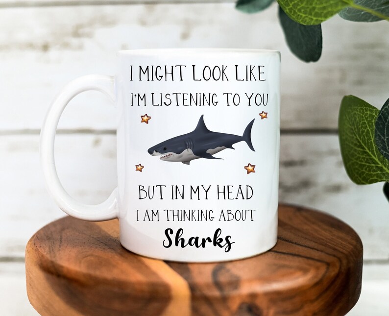 Pu&ograve; includere: Una tazza in ceramica bianca con un'illustrazione di squalo in bianco e nero e il testo "I might look like I'm listening to you but in my head I am thinking about Sharks".