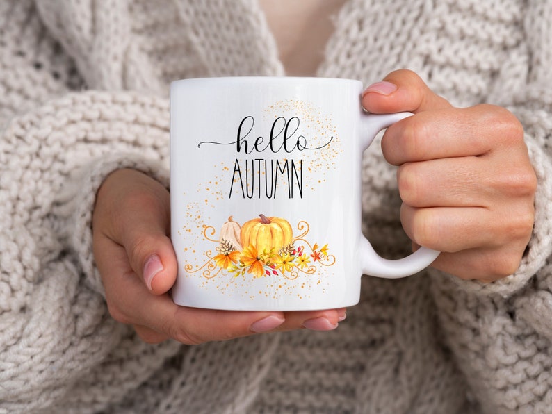 Puede incluir: Taza de cer&aacute;mica blanca con las palabras "hello AUTUMN" en letras negras cursivas y de imprenta. La taza presenta un dise&ntilde;o de acuarela de calabazas, follaje oto&ntilde;al y motas doradas. La taza es sostenida por una persona que lleva un su&eacute;ter color crema.