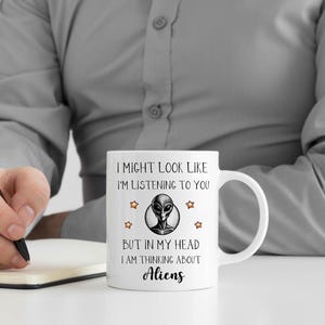 May include: White ceramic mug with a black and white alien graphic and the text "I might look like I'm listening to you, but in my head I am thinking about aliens."