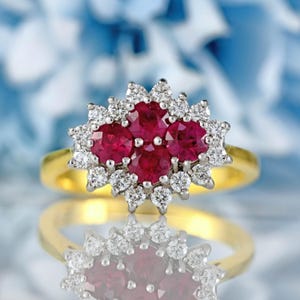 Peut inclure: Une bague en or ornée d'un groupe de cinq rubis rouges ronds, entourés d'un halo de petits diamants étincelants. La monture de la bague est en or poli, et les pierres précieuses sont serties sur une surface réfléchissante.
