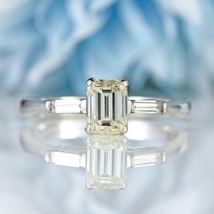 Peut inclure: Une bague en platine avec une grosse pierre centrale en diamant jaune clair de forme rectangulaire. La bague comporte deux diamants rectangulaires plus petits de chaque côté de la pierre centrale, sur fond bleu flou.