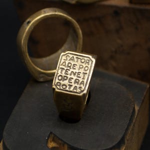 Può includere: Quattro anelli antichi color oro sono esposti su una superficie scura. Uno degli anelli presenta il testo "SATOR AREPO TENET OPERA ROTAS". Gli anelli sono posizionati su blocchi di legno, creando un'estetica vintage.