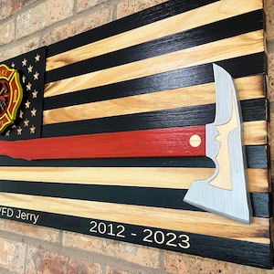 May include: A wooden wall plaque featuring a stylized American flag with black and natural wood stripes. A red-handled fire axe with a silver blade is mounted horizontally. A fire department emblem and the text BLVFD Jerry 2012 - 2023 are visible.