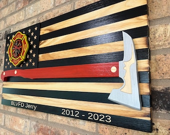 Puede incluir: Una bandera americana de madera con rayas negras y de madera natural. Una manguera de incendios roja está montada en la bandera, y un hacha de bombero plateada está montada en el lado derecho. La bandera tiene un emblema de bomberos amarillo y rojo en el lado izquierdo. El texto "BLVFD Jerry 2012-2023" está inscrito en la parte inferior de la bandera.