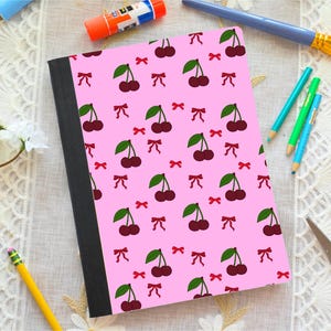 May include: A pink notebook with a pattern of cherries and red bows. The notebook has a black spine and is surrounded by art supplies, including pencils, a paintbrush, and glue.