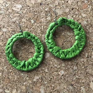 Könnte beinhalten: Ein Paar grüne Stoff-Creolen. Die Ohrringe sind aus gewebtem Stoff gefertigt und haben ein schlichtes, kreisrundes Design.